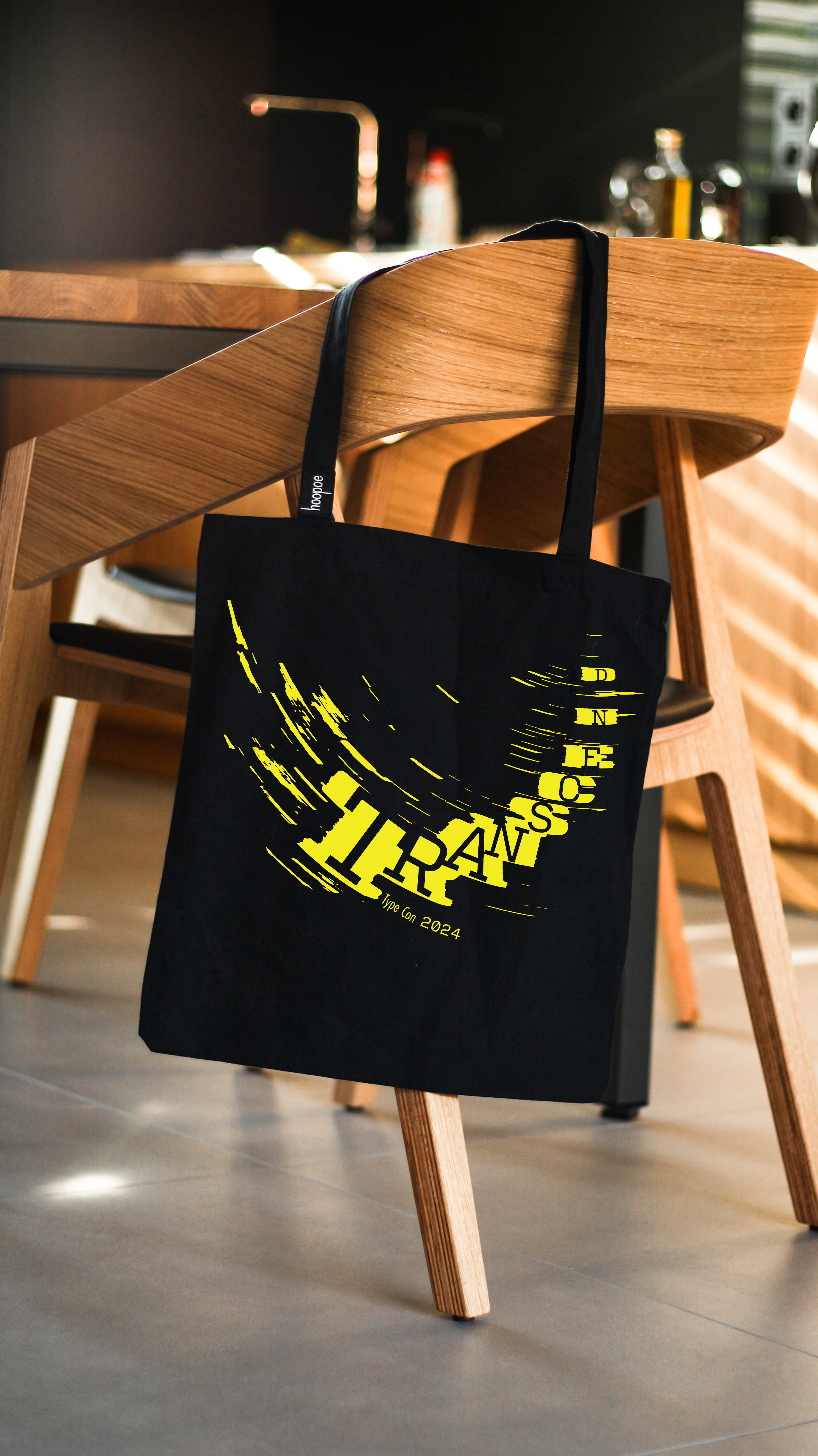 The transcend tote bag. its black with the yellow transcend logo. it hangs on a chair.